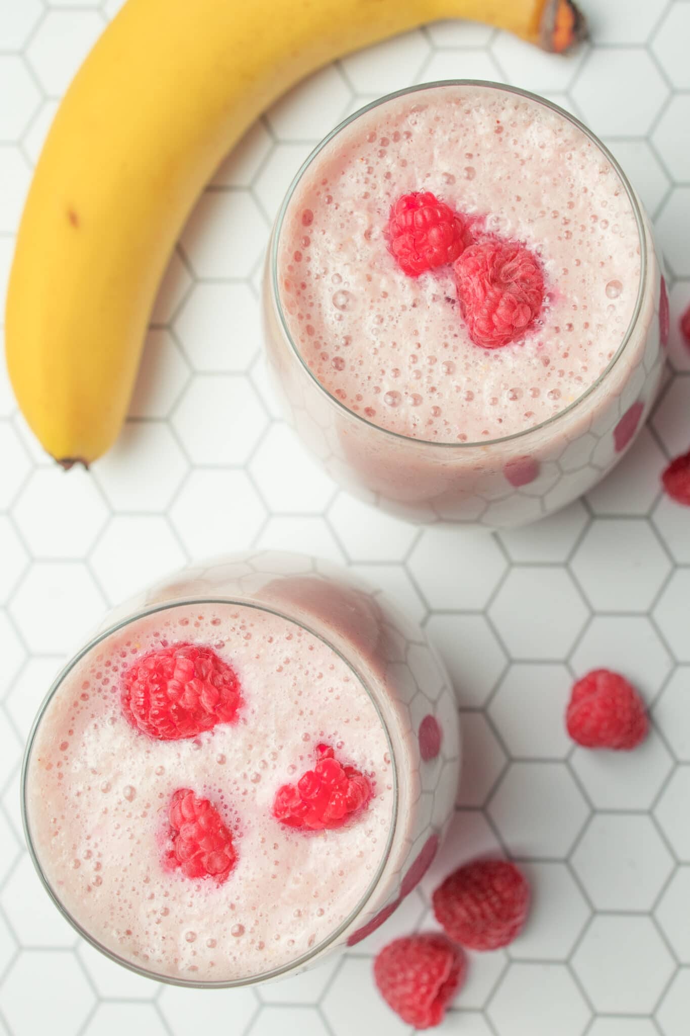 Two glasses of drink with a banana and raspberries surrounding them.