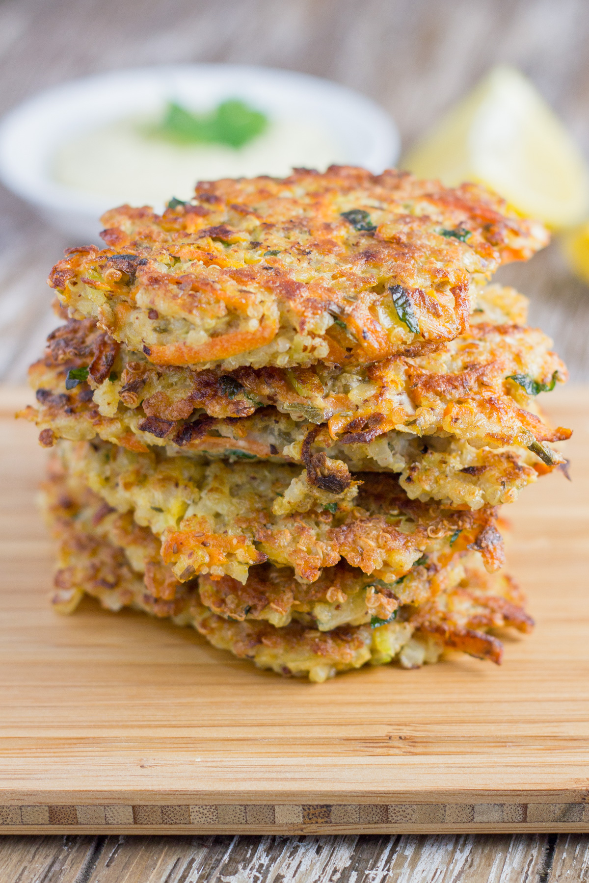 Stack of quinoa fritters on a board with garlic aioli and lemon wedges.