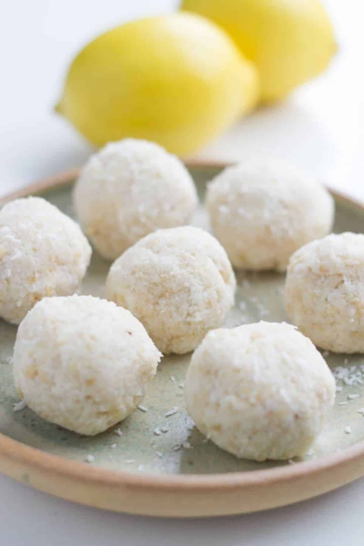 Bowl of lemon coconut bliss balls on a plate with some lemons behind them. 