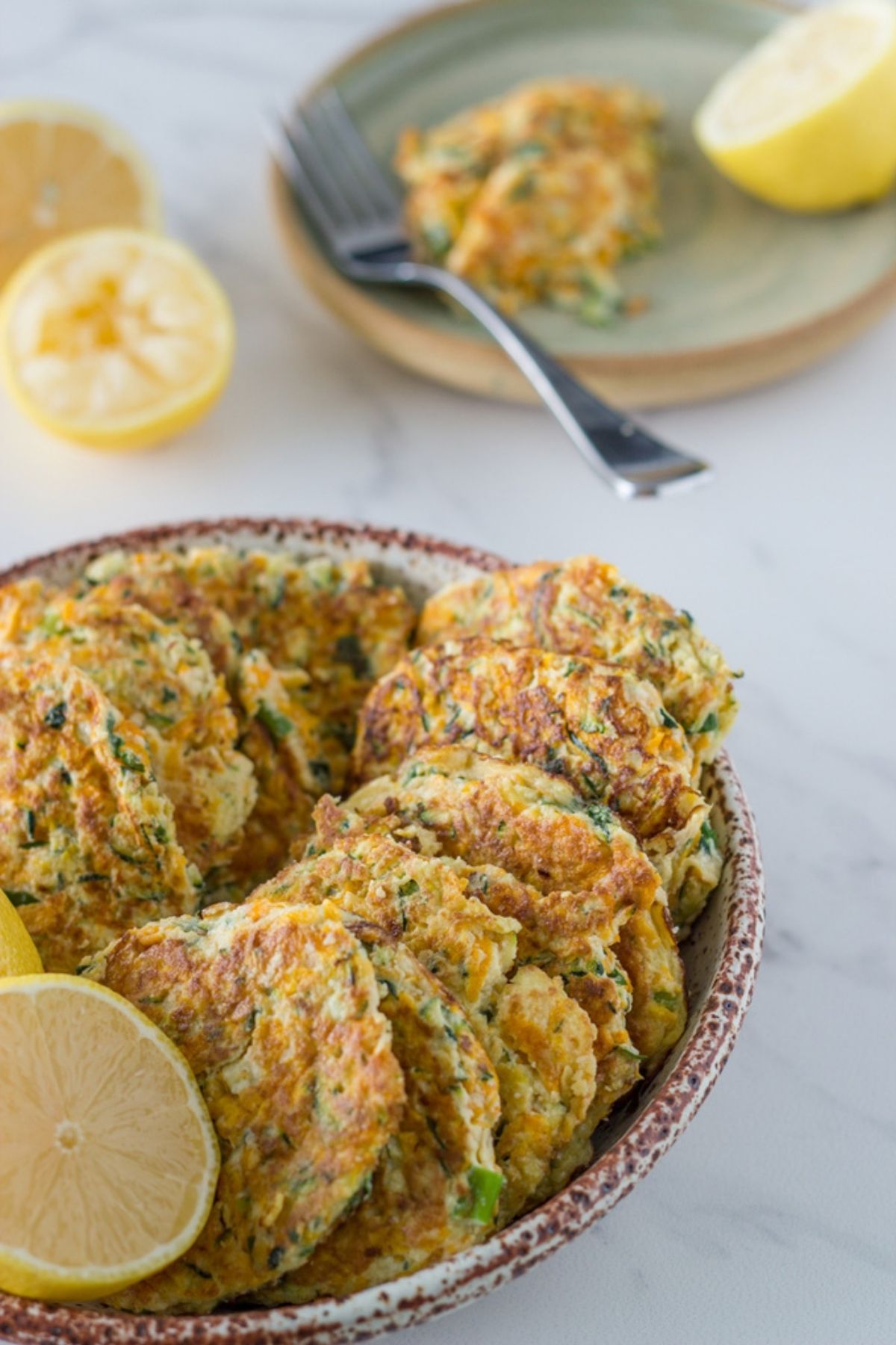 Bowl of pumpkin and zucchini fritters with some lemon and a place behind it with a fritter on it.