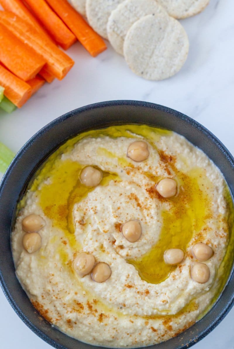 hummus in a black bowl topped with extra chick peas, olive oil and veggie sticks and crackers on the side