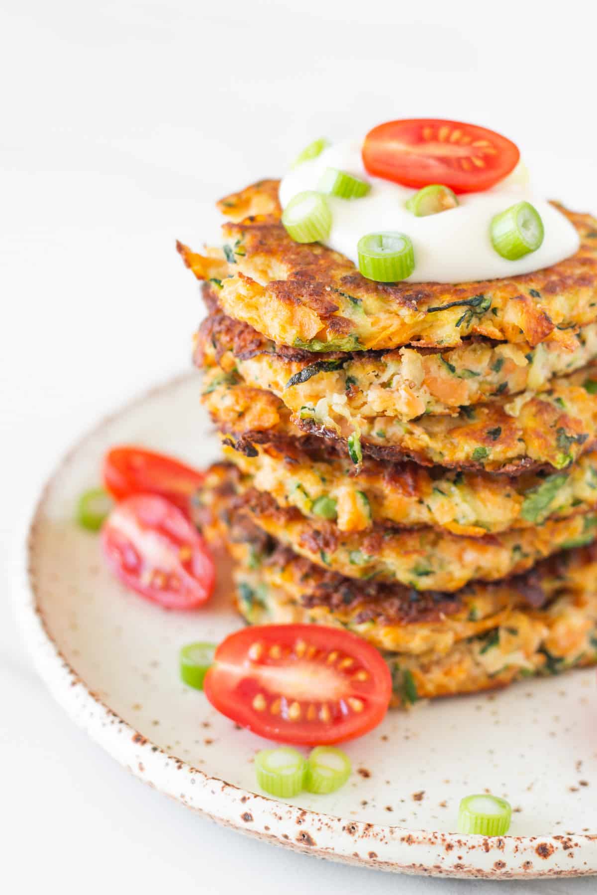 Healthy zucchini and sweet potato fritters stacked on a plate with aioli, cherry tomatoes and lemon wedges.