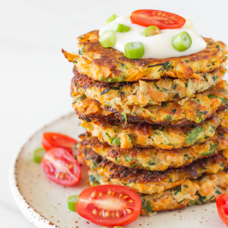 stack healthy zucchini and sweet potato fritters on a plate with aioli, cherry tomatoes and lemon wedges.