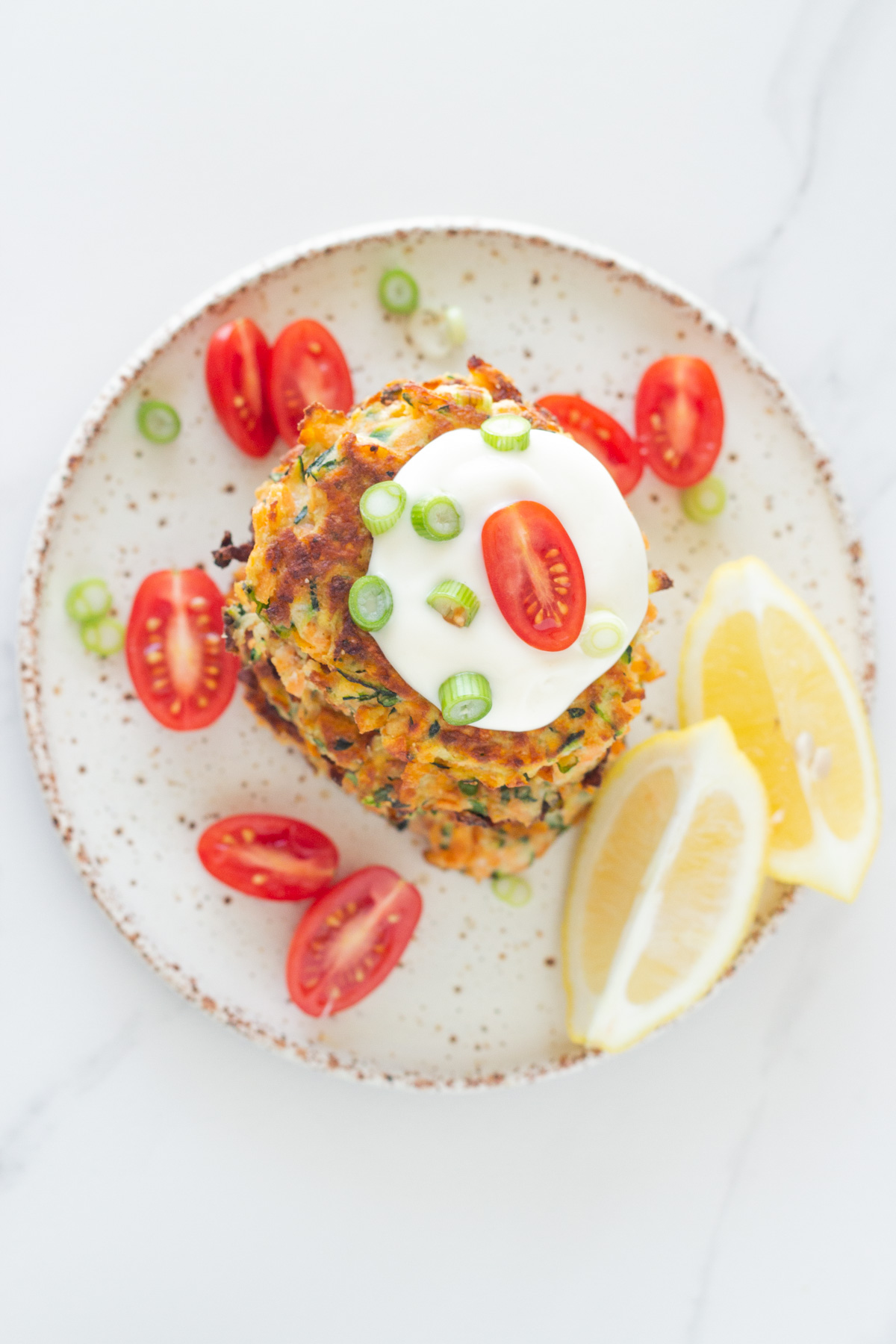 top down view of a stack of the cooked fritters on a plate with aioli, cherry tomatoes and lemon wedges.