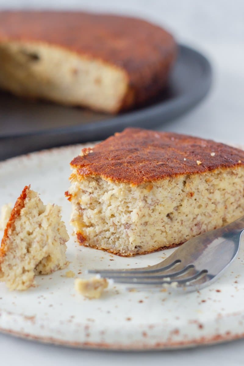 A slice of gluten free banana cake on a plate with the rest of the cake behind it.