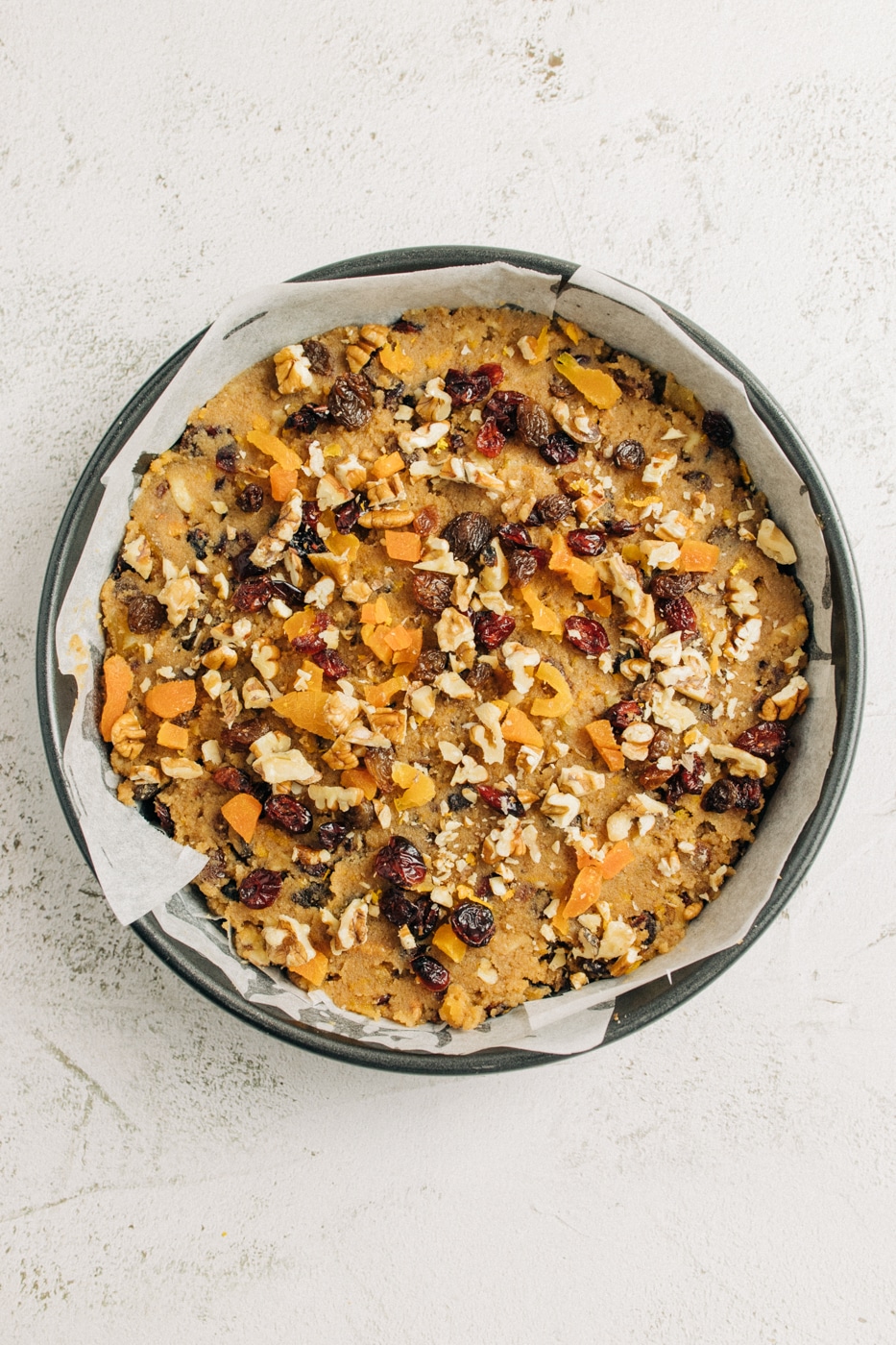 Cake batter in a lined cake tin, with added nuts and fruit on top, ready to be baked.