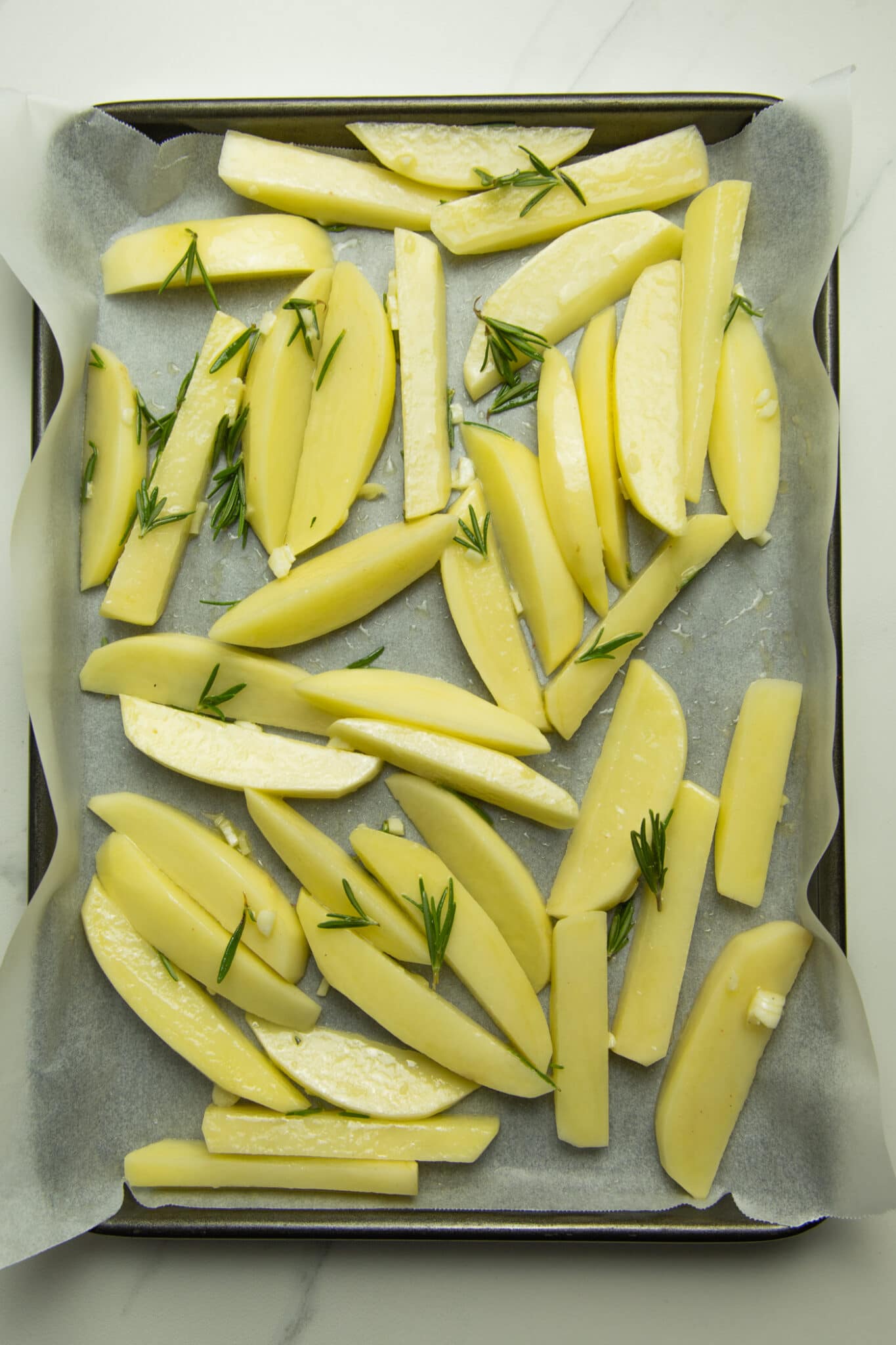 photo of uncooked fries on a baking tray lined with baking paper, topped with fresh garlic, rosemary and sea salt.