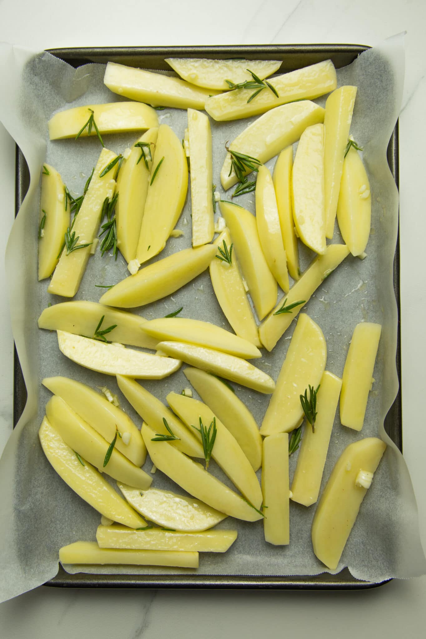  photo of uncooked fries on a baking tray lined with baking paper, topped with fresh garlic, rosemary and sea salt.