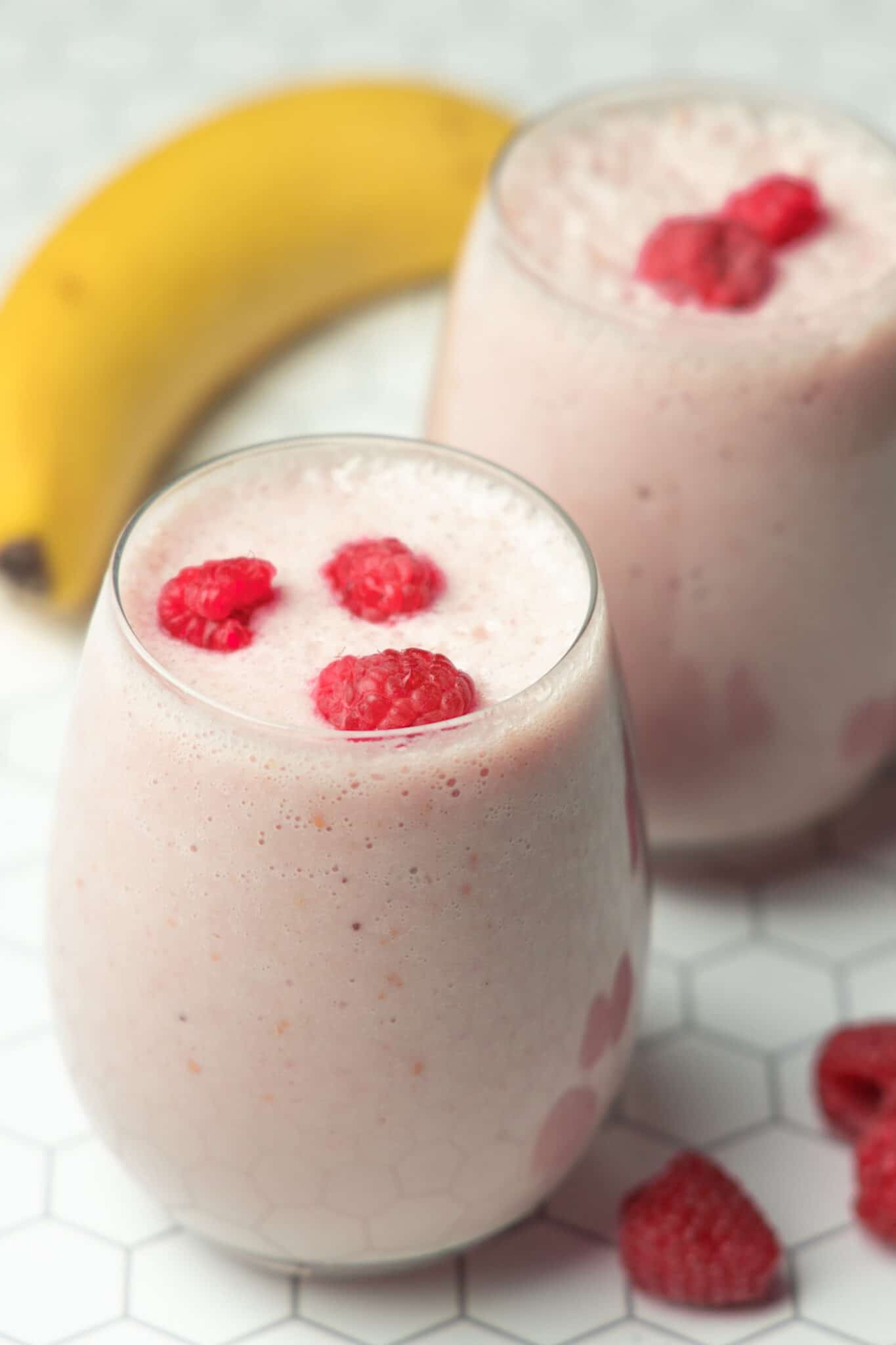 Two glasses of raspberry banana smoothie on a white tiled background.