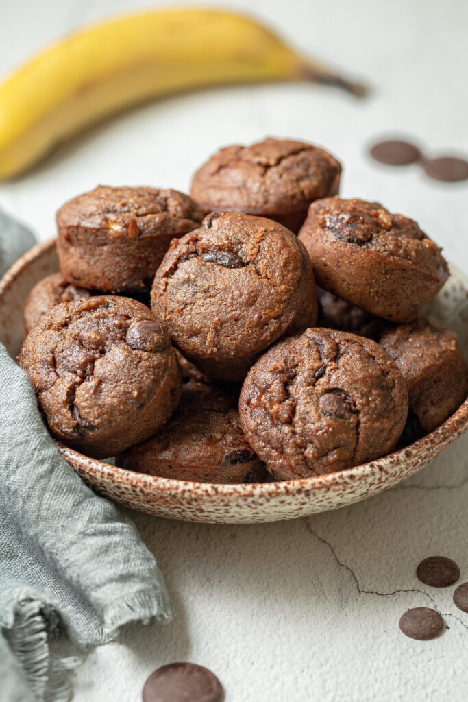 photo of a Bowl of Gluten Free Double Chocolate Banana Muffins next to a napkin, some chocolate chips and a banana in the background