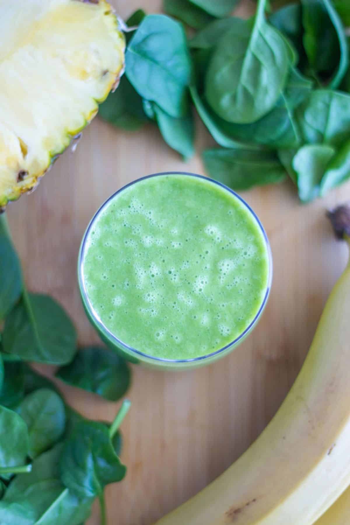 A glass of banana pineapple smoothie with pineapple. banana and bay spinach surrounding it.