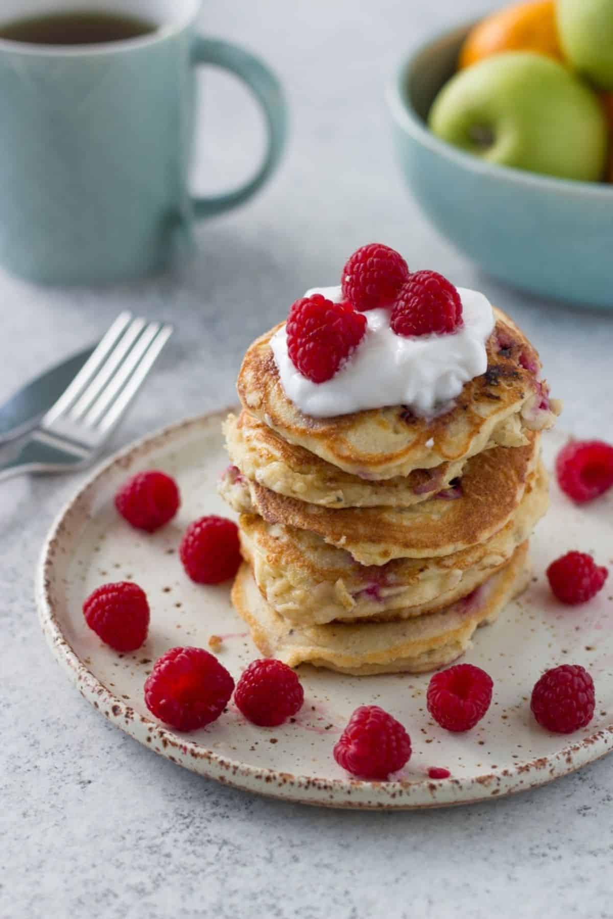 A big stack of raspberry pancakes on a plate with a cup of tea and bowl of fruit behind it.