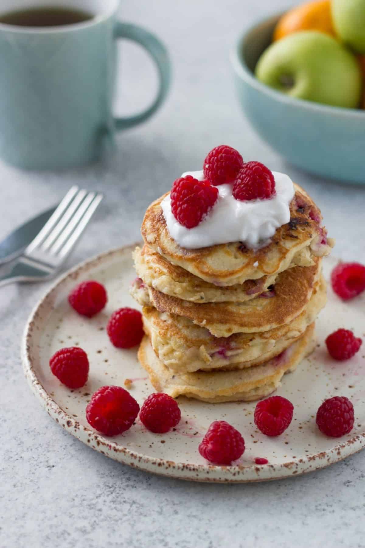 A big stack of raspberry pancakes on a plate with a cup of tea and bowl of fruit behind it. 